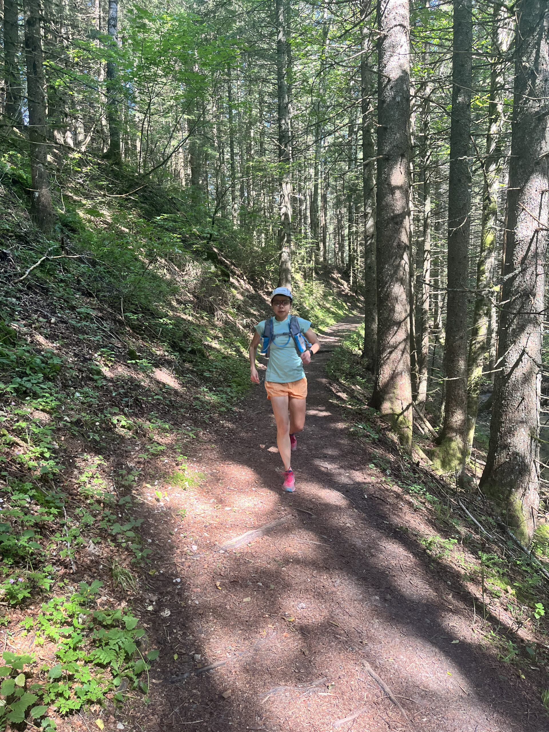 A runner in a cap, sunglasses, and orange shorts jogging along a shaded dirt trail through a pine forest near Villard-de-Lans.